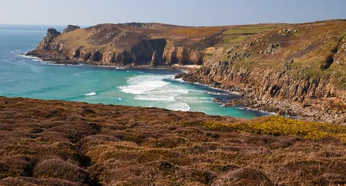 This landscape photograph shows the Landsend cliffs on the coast of Cornwall in the United Kingdom. Taken in the early afternoon during the spring season, the image captures the rugged natural beauty of the Cornish coastline, with dramatic cliffs rising sharply from the turquoise waters of the Atlantic Ocean. The foreground features brown and green vegetation typical of springtime coastal nature, while the background highlights the impressive rocky formations and gentle slopes of the cliffs. The composition showcases the raw texture and geological features that define this iconic part of Cornwall’s coast.